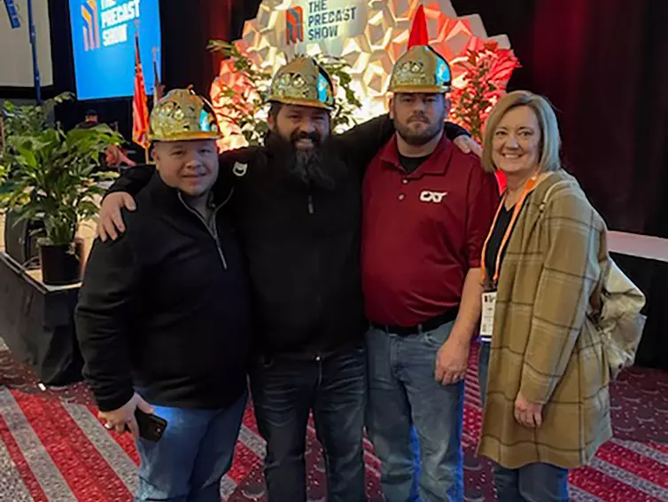 three men in gold hard hats and a woman smiling in front of a stage at the Precast Show.