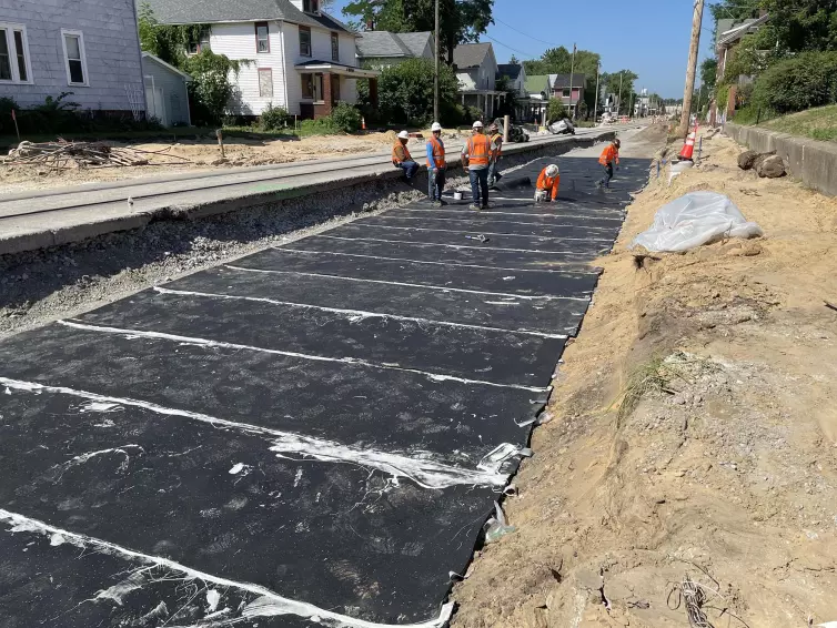 Six workers in hard hats and orange vests installing a ballast mat next to a train track with houses in the background.