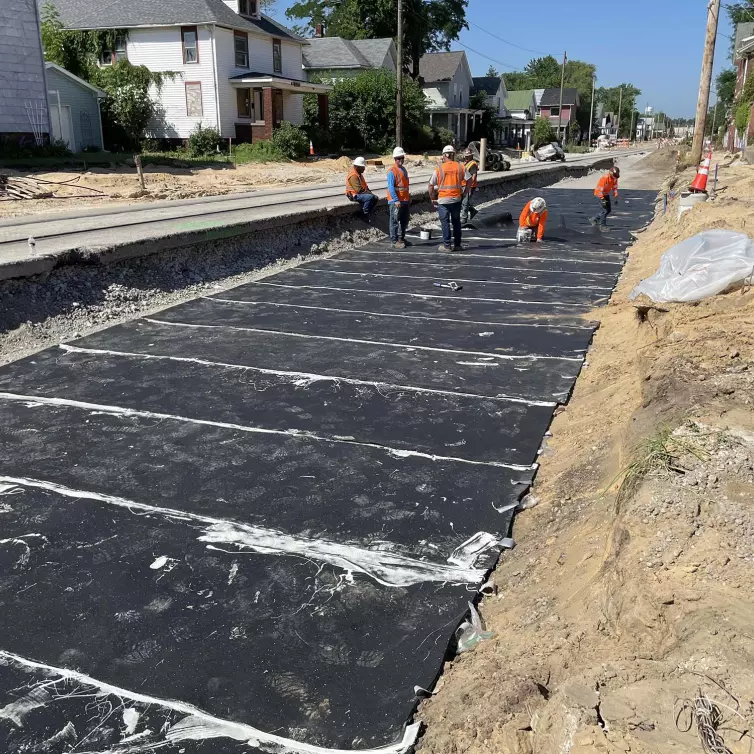 Six workers in hard hats and orange vests installing a ballast mat next to a train track with houses in the background.