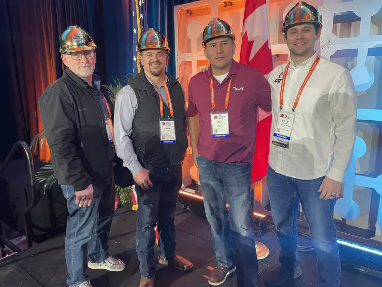 Four men smiling on a stage wearing gold hard hats.