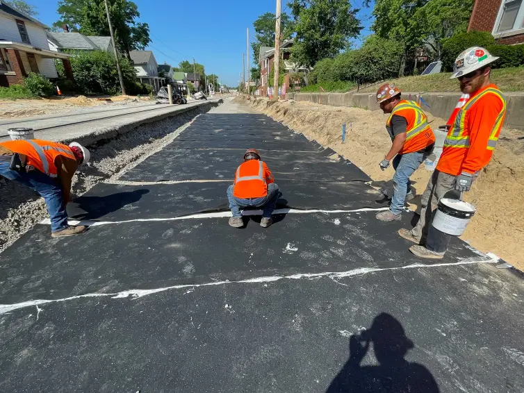 Four workers in hard hats and orange vests installing a ballast mat with houses in the background.