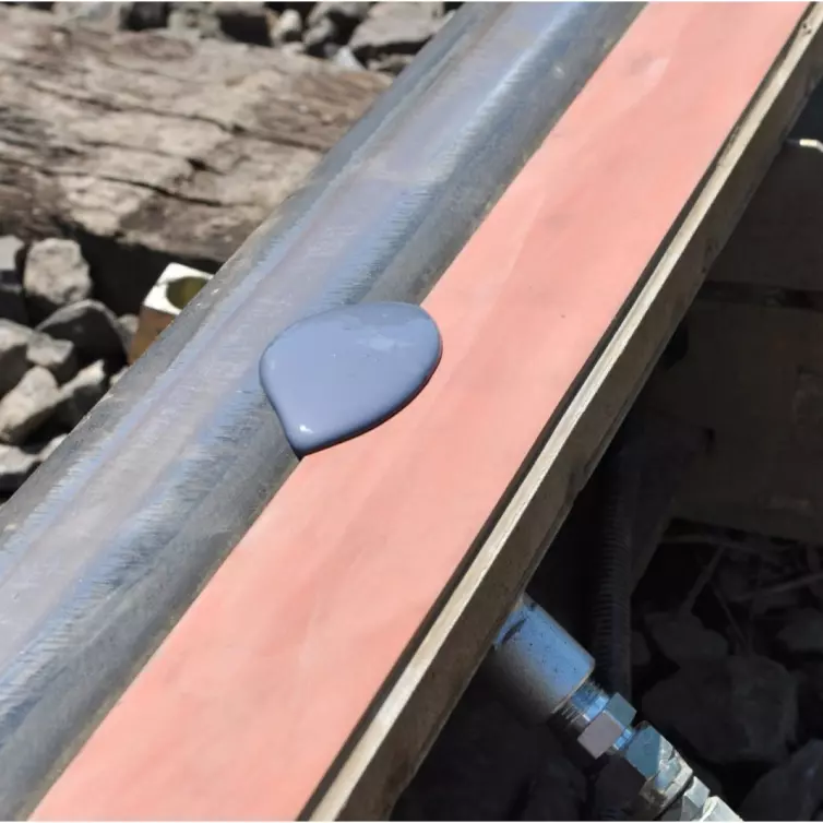 A close-up view of a railroad track where a puddle of purple-grey liquid is shown on an orange foam  bar attached to the rail.