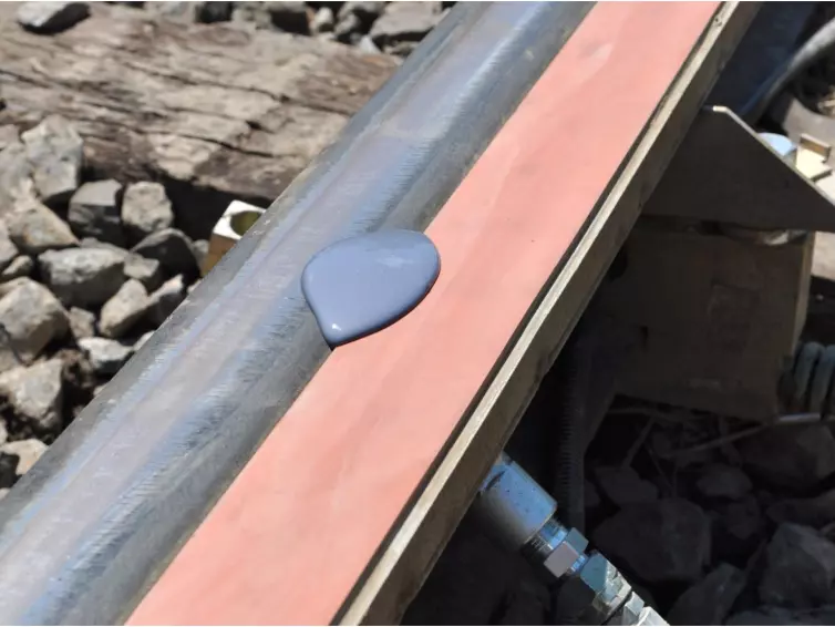 A close-up view of a railroad track where a puddle of purple-grey liquid is shown on an orange foam  bar attached to the rail.