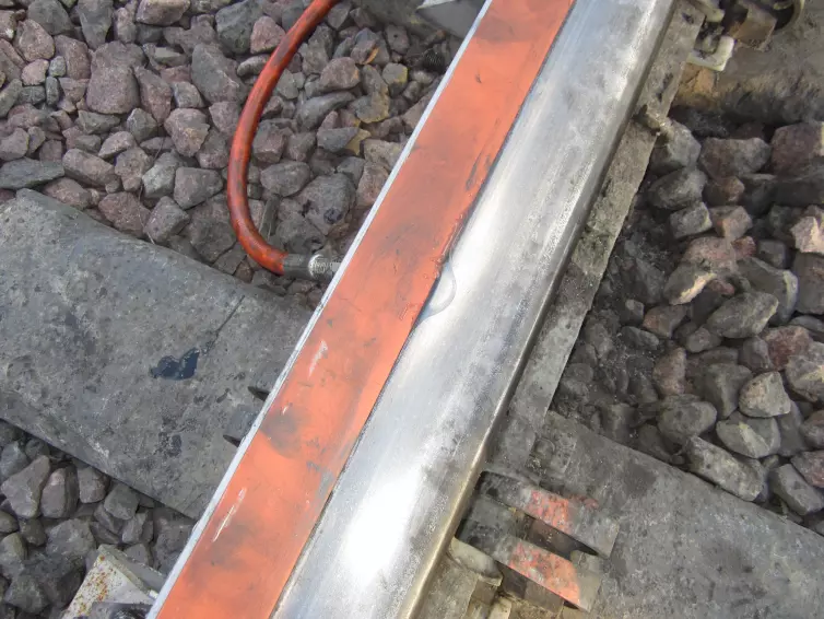 A close-up view of a railroad track where a puddle of clear  liquid is shown on an orange foam  bar attached to the rail.