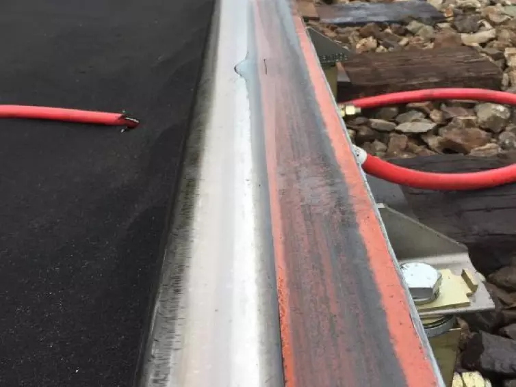 A close-up view of a railroad track where a puddle of clear  liquid is shown on an orange foam  bar attached to the rail.