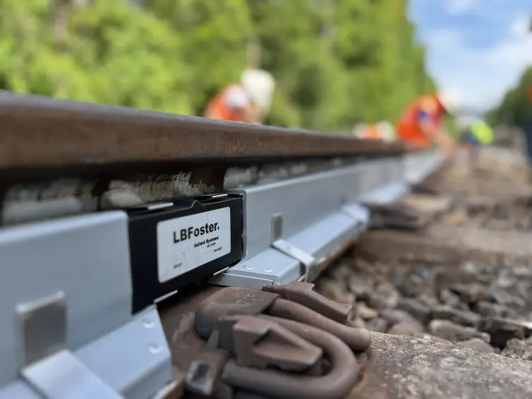 Close-up of L.B. Foster branded WILD IV strain gauge sensor attached to railway track with cables, showing railway workers in high-visibility clothing working in the blurred background.