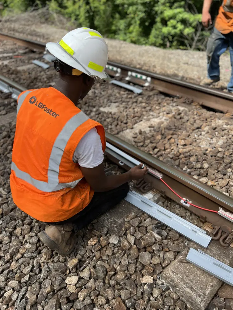 L.B. Foster technician in orange high-visibility safety vest and white hard hat kneeling beside railway tracks, installing strain gauge monitoring equipment with visible cables and circuit boards on the rail.