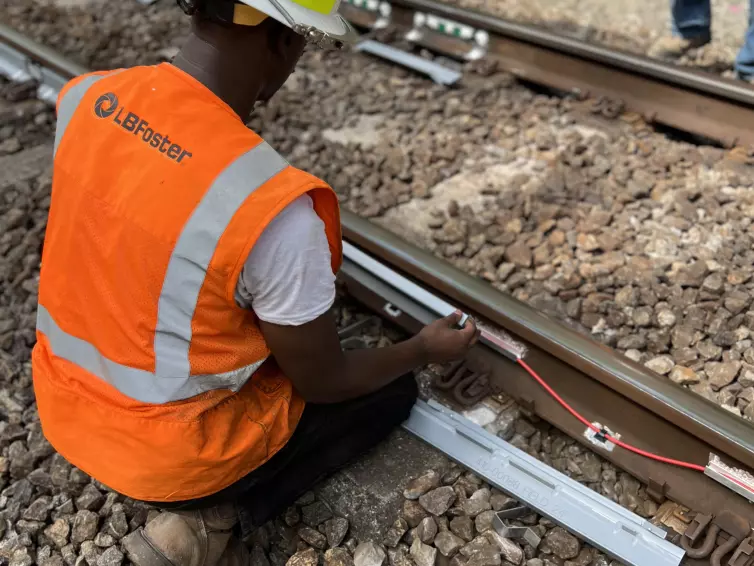 L.B. Foster technician in orange high-visibility safety vest and white hard hat kneeling beside railway tracks, installing strain gauge monitoring equipment with visible cables and circuit boards on the rail.