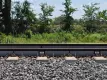 Railway tracks with L.B. Foster branded strain gauge sensors mounted on the rails between concrete sleepers, surrounded by ballast stones, with lush green vegetation in the background.