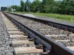 Railroad tracks with WILD IV monitoring sensors attached to the rails, surrounded by ballast stones and wooden sleepers, with signal equipment and green vegetation visible in the background under a partly cloudy sky.