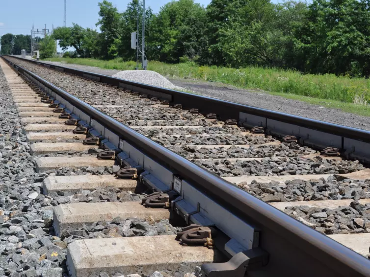 Railroad tracks with WILD IV monitoring sensors attached to the rails, surrounded by ballast stones and wooden sleepers, with signal equipment and green vegetation visible in the background under a partly cloudy sky.