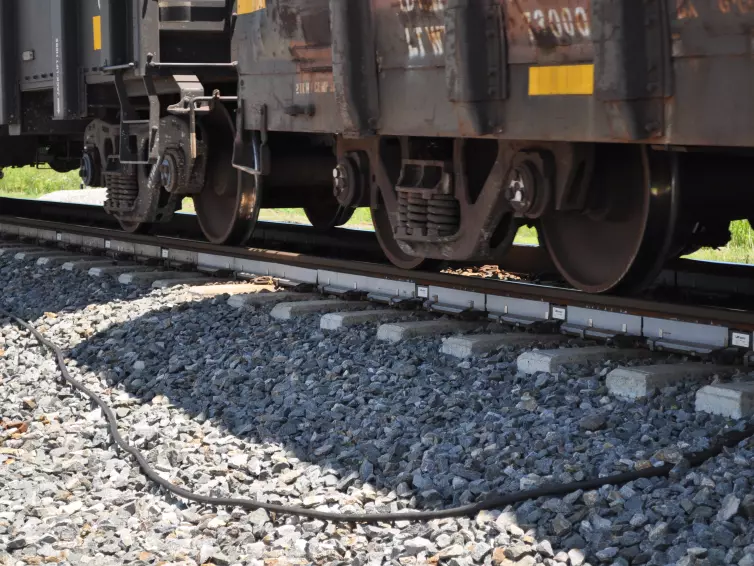 Freight train wheels and bogies passing over railway track equipped with WILD IV monitoring sensors on concrete sleepers, with ballast stones and connecting cables visible beneath the train.