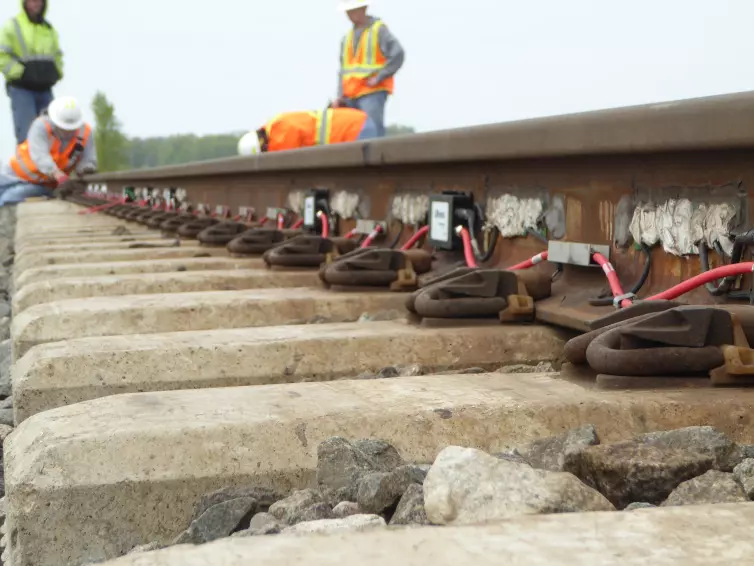 Railway workers in high-visibility safety gear installing WILD IV monitoring equipment on railroad tracks, with multiple L.B. Foster sensor units and red cables visible along the rail sections, concrete sleepers and ballast stones in foreground.